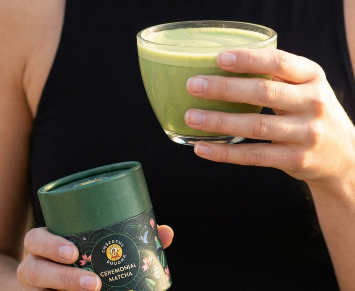 A hand holding a bright green matcha latte beside the Cheerful Buddha Ceremonial Matcha tube