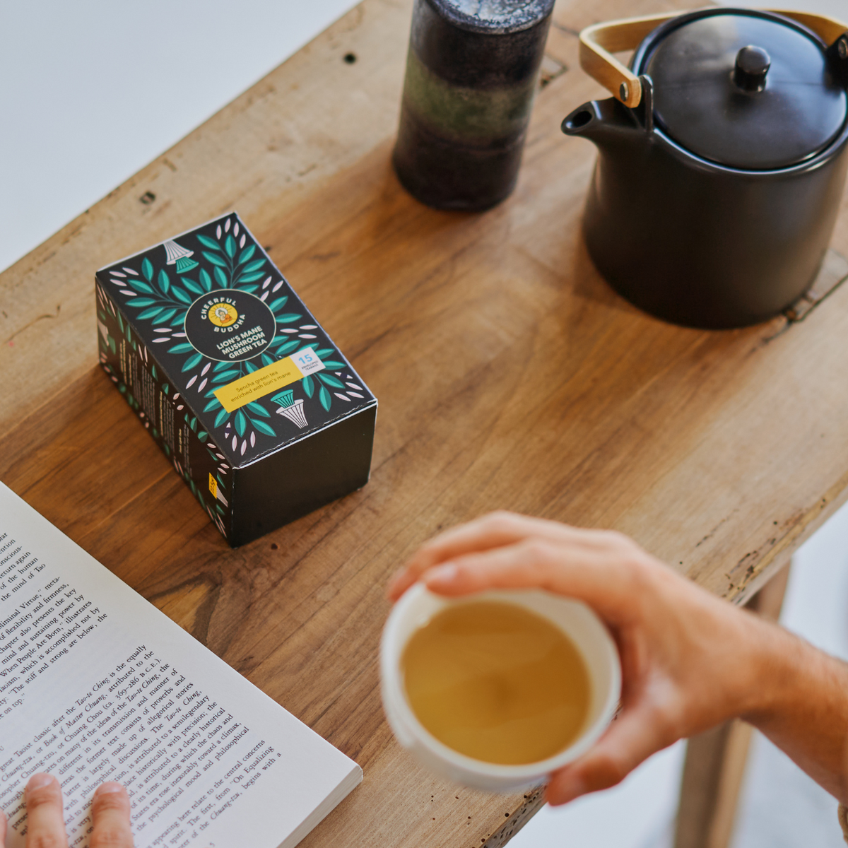 Lion's Mane Green Tea box on a wooden table with a teapot, an open book and a cup of green tea