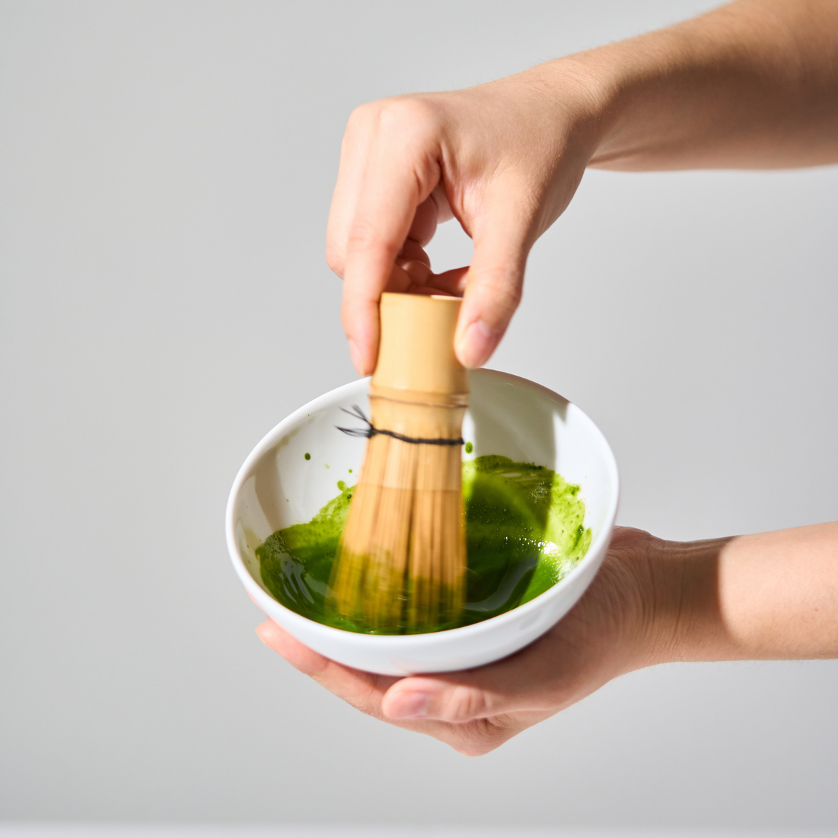 Hands whisking ceremonial matcha in a white bowl with a bamboo whisk